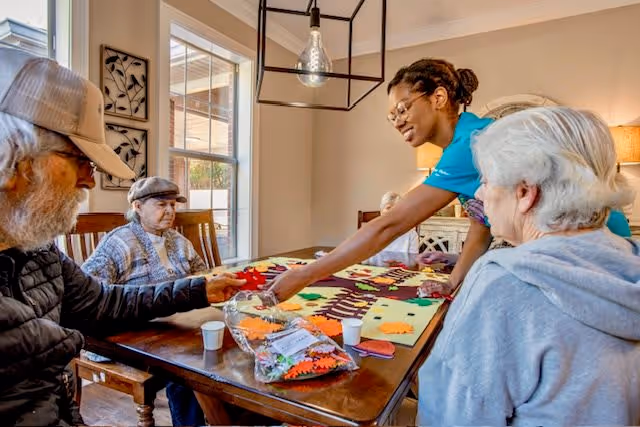 A group of elderly people sitting around a wooden table engaged in a craft activity with colorful felt pieces, assisted by a caregiver in a bright room with large windows and a hanging light fixture.