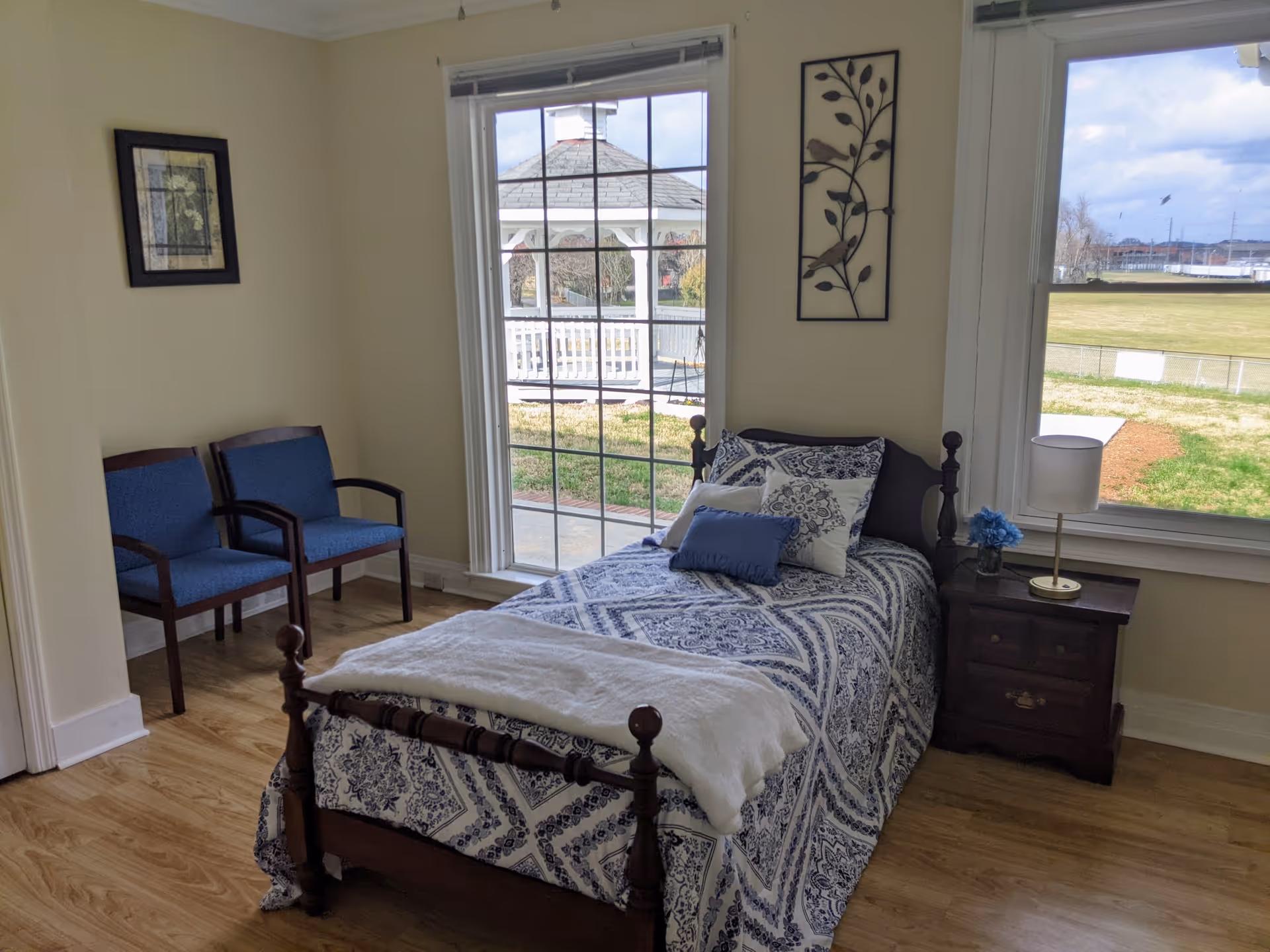 A small bedroom with a single wooden bed covered in a blue and white patterned bedspread and several pillows. Next to the bed is a wooden nightstand with a lamp and a small vase of blue flowers. Two blue upholstered chairs are placed against the wall. Large windows provide a view of a gazebo and a grassy outdoor area.