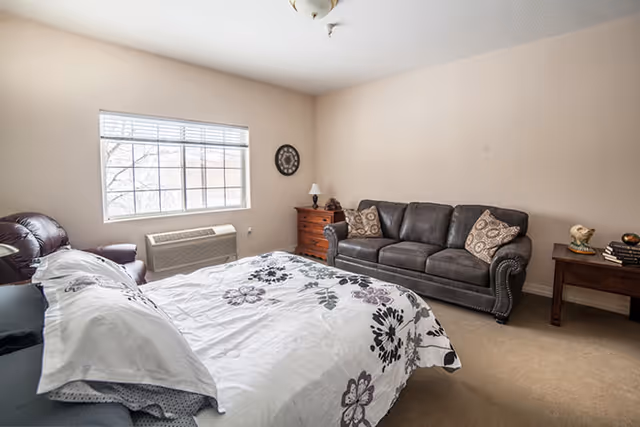 Bright furnished bedroom with a bed, upholstered sofa, nightstands, and a large window.