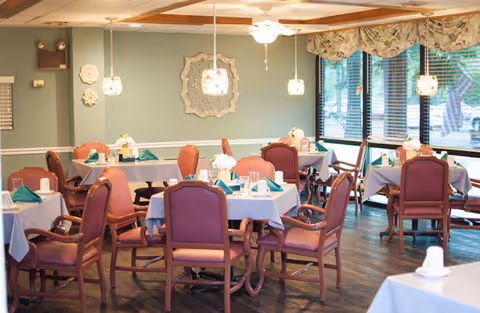 A dining room with multiple tables covered in light blue tablecloths, each set with green folded napkins, white flowers in small vases, and glassware. The room features wooden chairs with purple cushions, pendant lights hanging from the ceiling, large windows with floral valances, and a decorative wall piece.