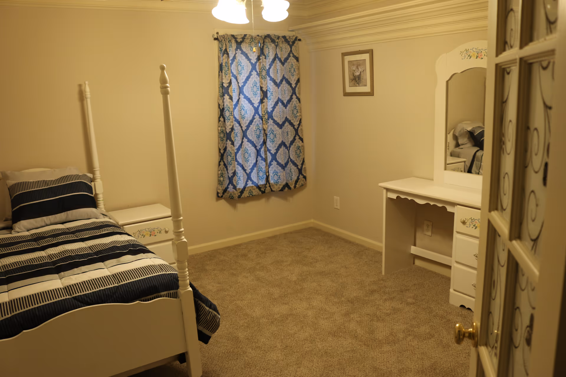 A small bedroom with beige walls and carpet. The room contains a white four-poster bed with striped bedding, a white nightstand with floral details, a window with blue and white patterned curtains, a white vanity desk with a mirror, and a framed picture on the wall.