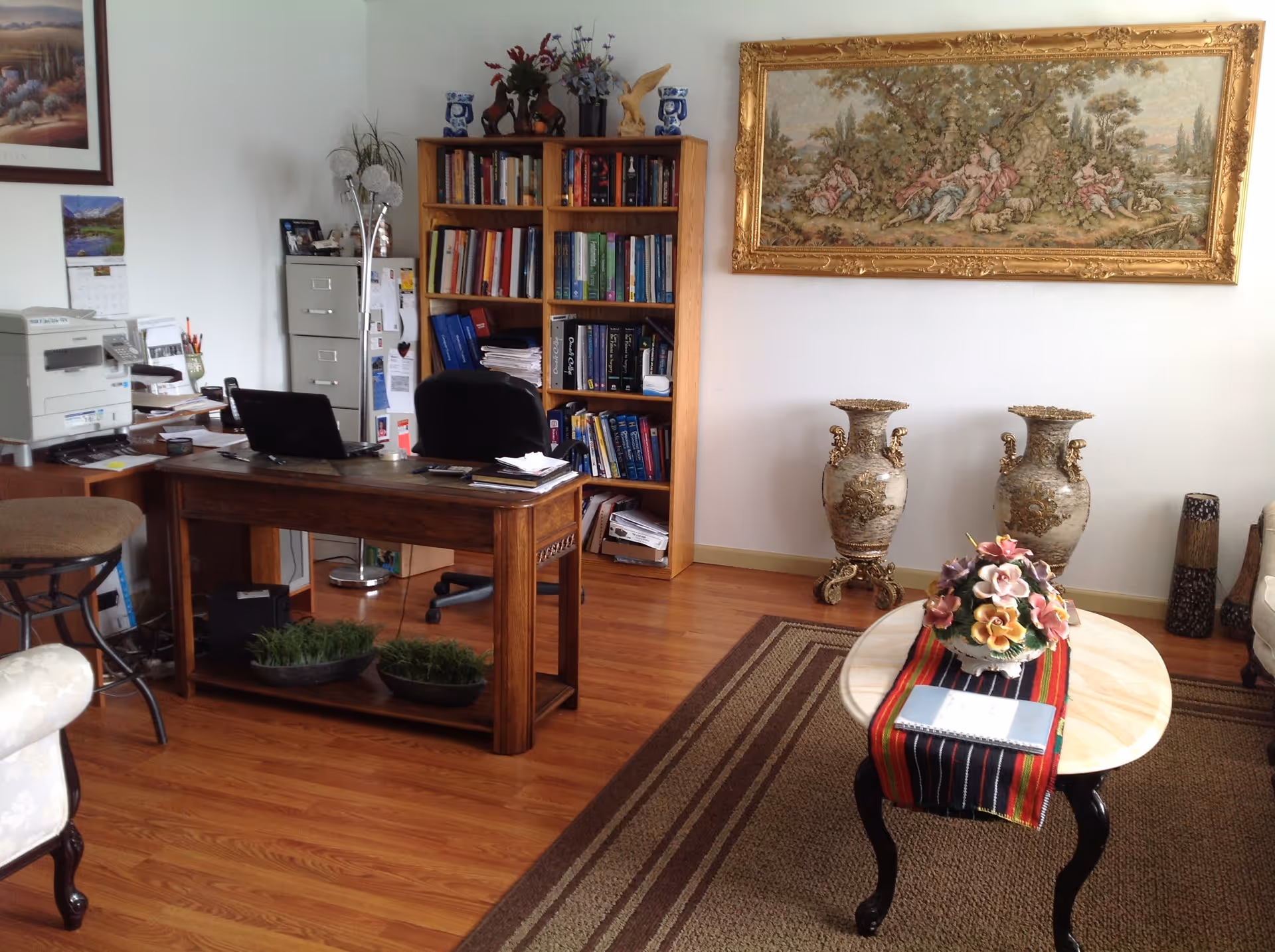 A combined living room and home office with a wooden desk and chairs, a bookshelf, a round coffee table with a floral arrangement, and two ornate vases against the wall.