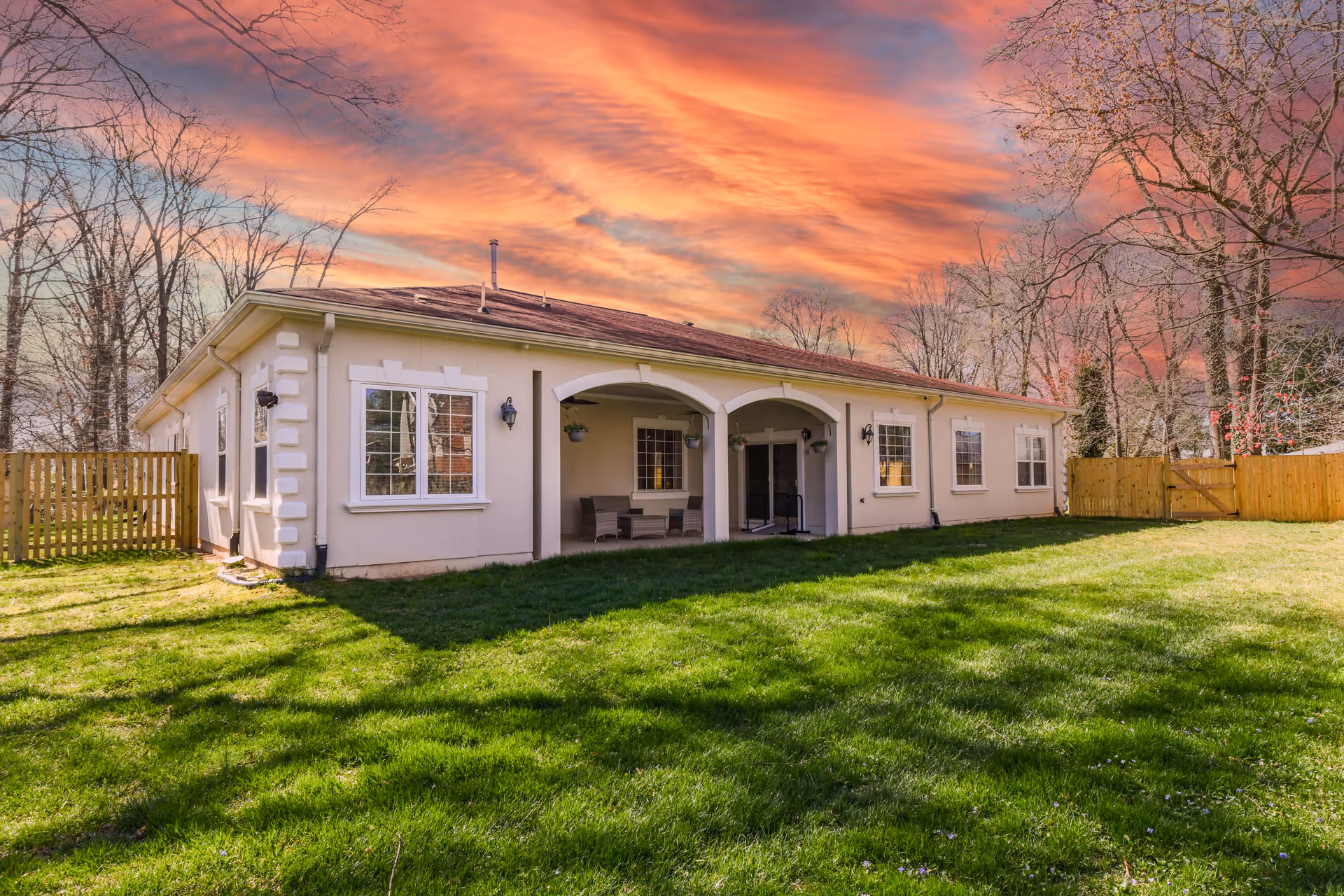Single-story light-colored building with a covered patio and chairs opening onto a green lawn beneath a dramatic sunset sky.