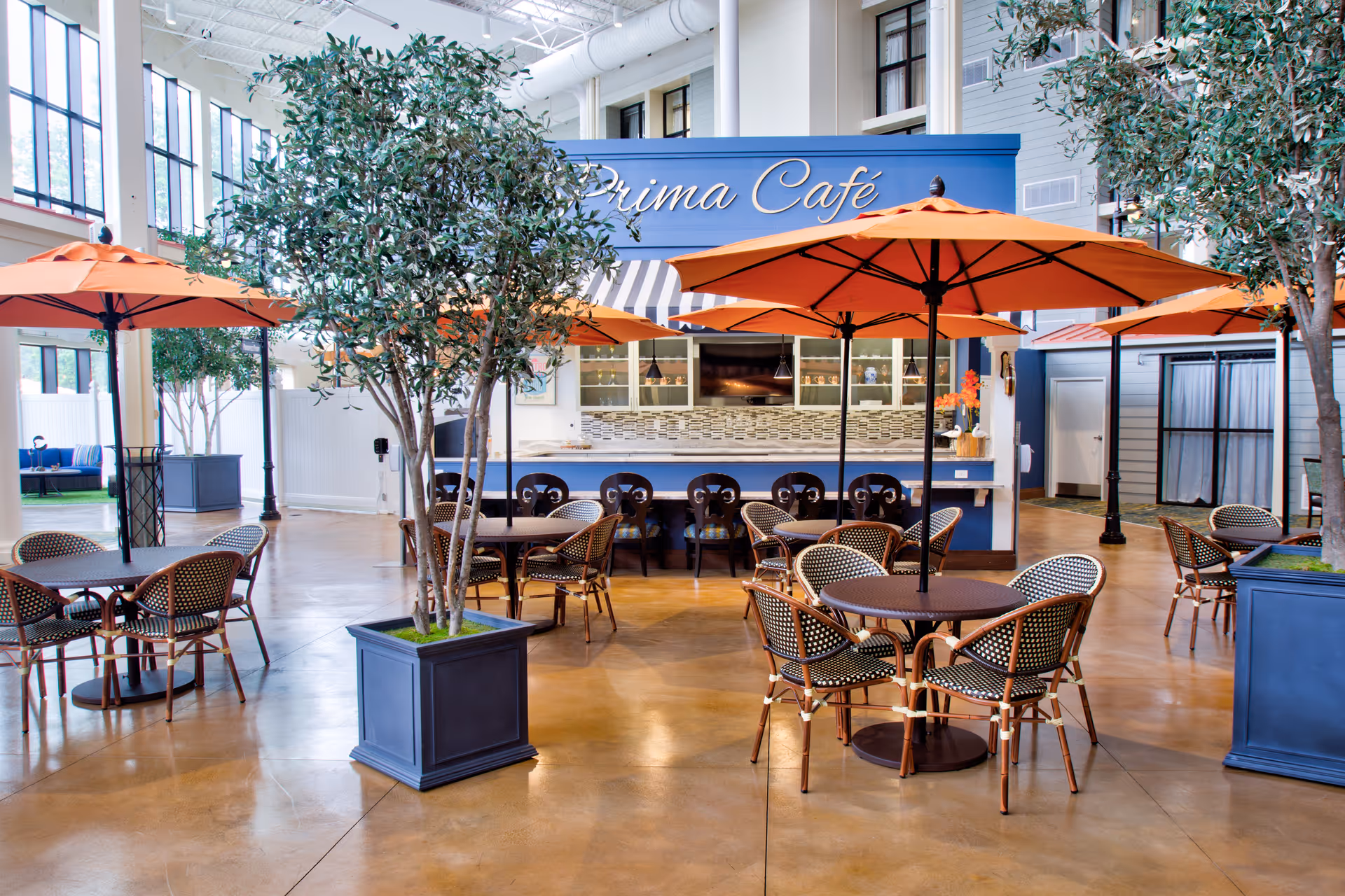 Indoor café area with several round tables and chairs, each table shaded by large orange umbrellas. There are tall potted trees placed around the space. In the background, a counter with bar stools and a sign reading 'Prima Café' is visible. The area has large windows letting in natural light and a polished floor.