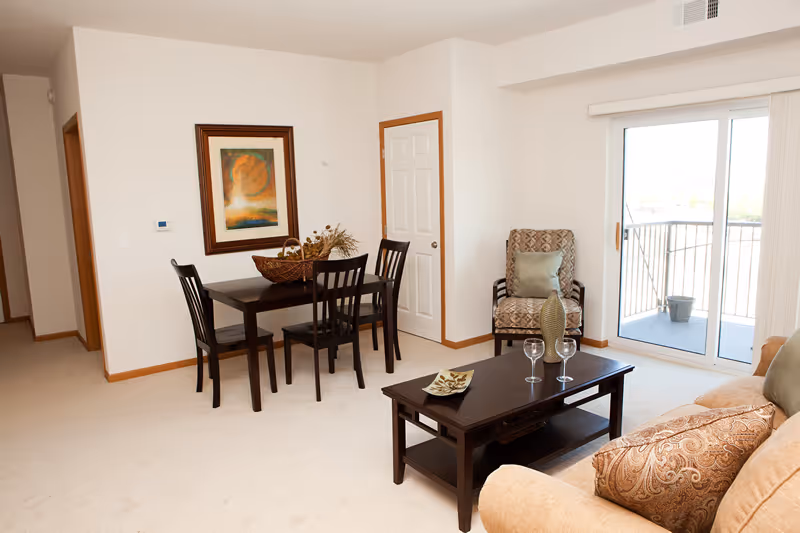 Open living room and dining area with a dark wood dining table, coffee table, sofa and armchair, and a sliding glass door to a balcony.