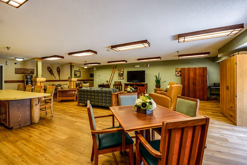 A spacious senior living common area with wooden floors and multiple seating arrangements including a table with four chairs and a flower centerpiece, sofas, and armchairs. The room features green walls decorated with vintage snowshoes and framed pictures, a flat-screen TV on a wooden stand, and warm ceiling lights.