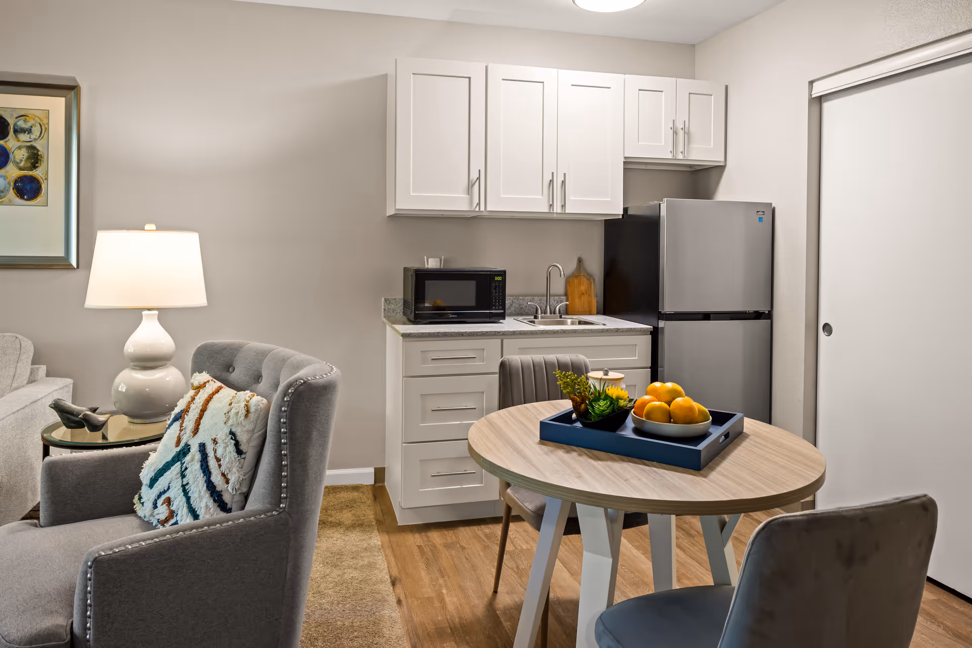 A small kitchen area with white cabinets, a microwave, a sink, and a stainless steel refrigerator. In front of the kitchen is a round wooden table with two chairs, decorated with a tray holding a bowl of oranges and a small plant. To the left, there is a gray armchair with a colorful textured pillow and a side table with a white lamp. The room has light-colored walls and wood flooring.