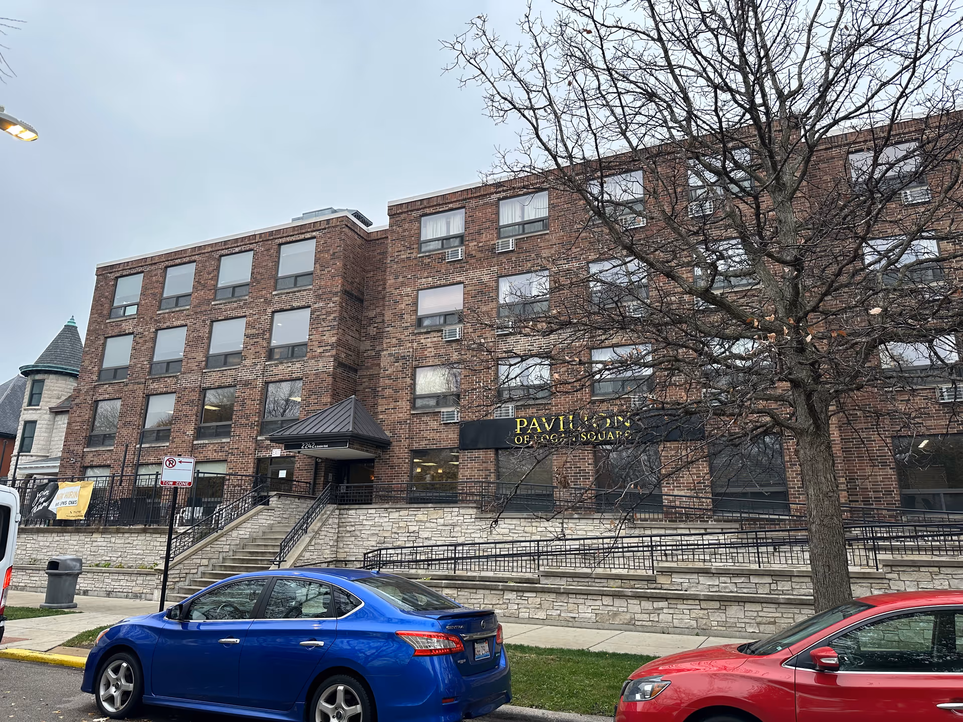 Exterior view of a multi-story brick building with a sign that reads Pavilion of Logan Square. There are several windows with air conditioning units, a tree without leaves in front, and two parked cars, one blue and one red, along the street.