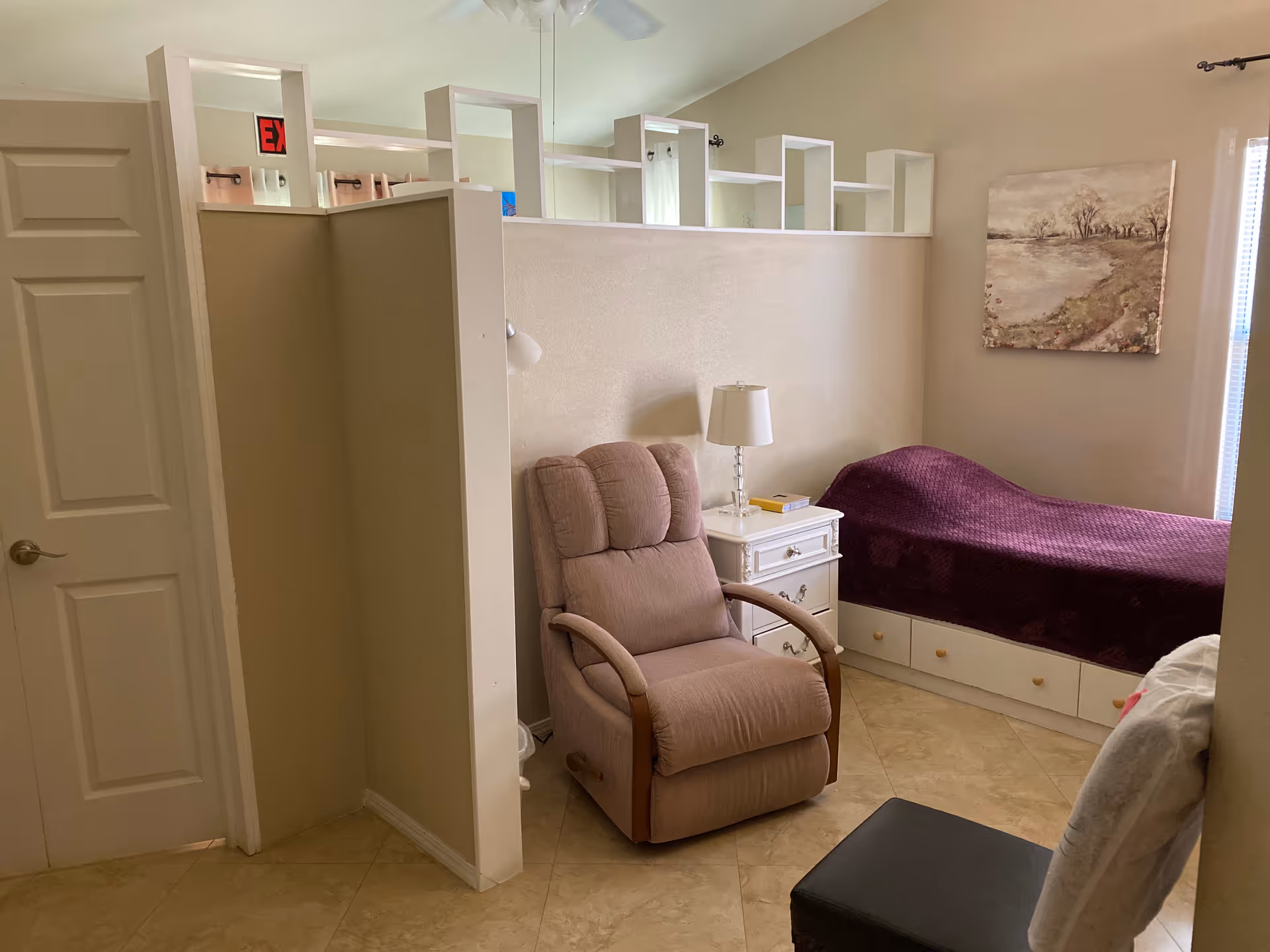 A beige recliner chair beside a white nightstand and a single bed with a purple cover.