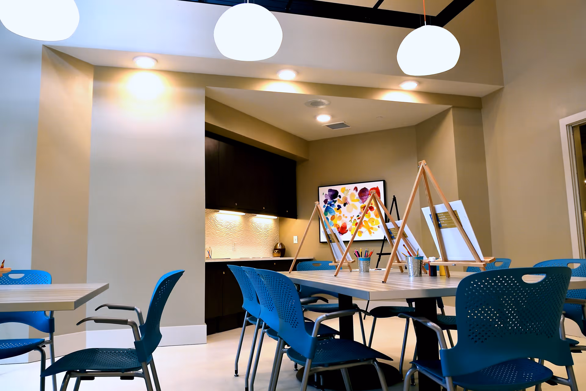 Bright interior activity room with tables, blue chairs, easels and art supplies under hanging lights.