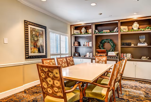 A well-lit dining room with a rectangular table surrounded by eight patterned chairs. The room features a large built-in wooden bookshelf filled with decorative items, books, and greenery. A window with white shutters and a framed abstract painting are visible on one wall.