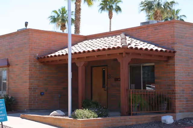 Entrance of a low brick building with a tiled porch roof, flagpole, doorway, and palm trees in the background.