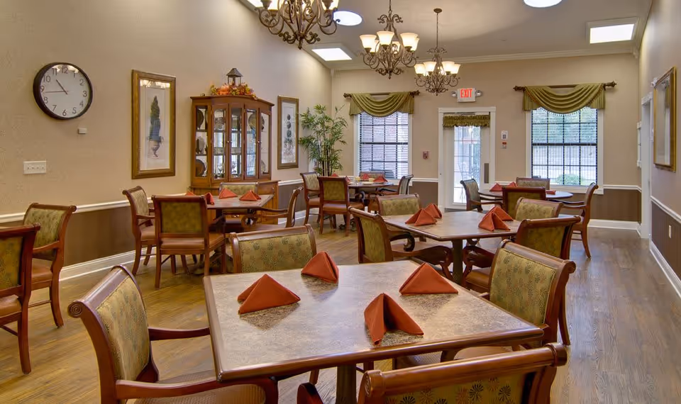 A dining room in a senior living facility with multiple wooden tables and chairs. Each table has four chairs and is set with folded red napkins. The room features wooden flooring, beige walls with white trim, framed artwork, a glass-front cabinet, two windows with green valances, and two chandeliers hanging from the ceiling. There is an exit door at the far end of the room.