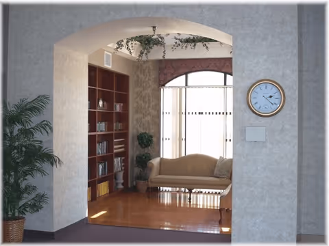 Sunlit seating area with a couch, bookshelf, plants and a wall clock viewed through an arched doorway.
