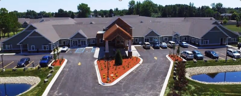 Front exterior of a single-story senior living building with a covered entrance, parking lot, landscaped islands, and small ponds.