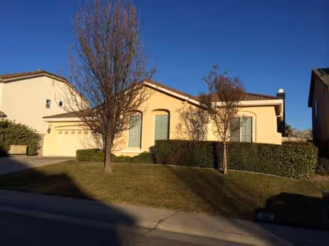 Front exterior of a single-story beige senior living facility with trees and hedges.