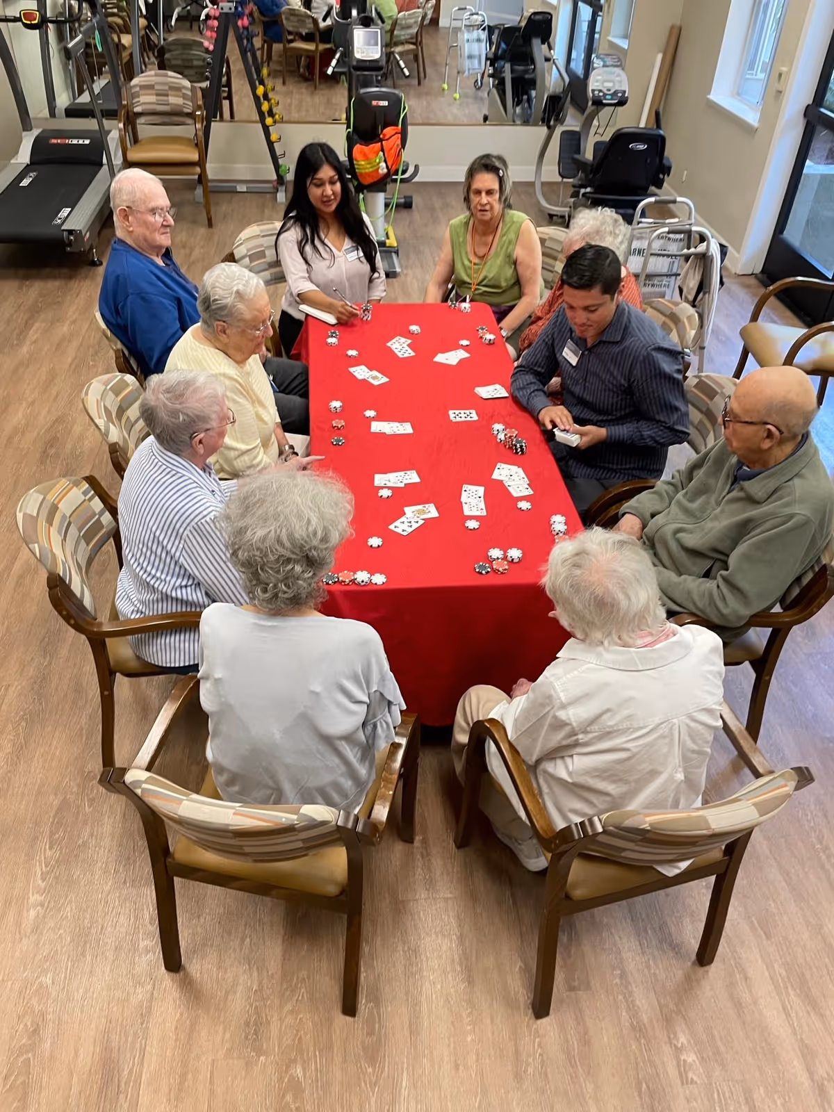 A group of elderly people and two younger adults sitting around a rectangular table with a red tablecloth, playing a card game with poker chips. The room has wooden flooring, exercise equipment, and chairs in the background.