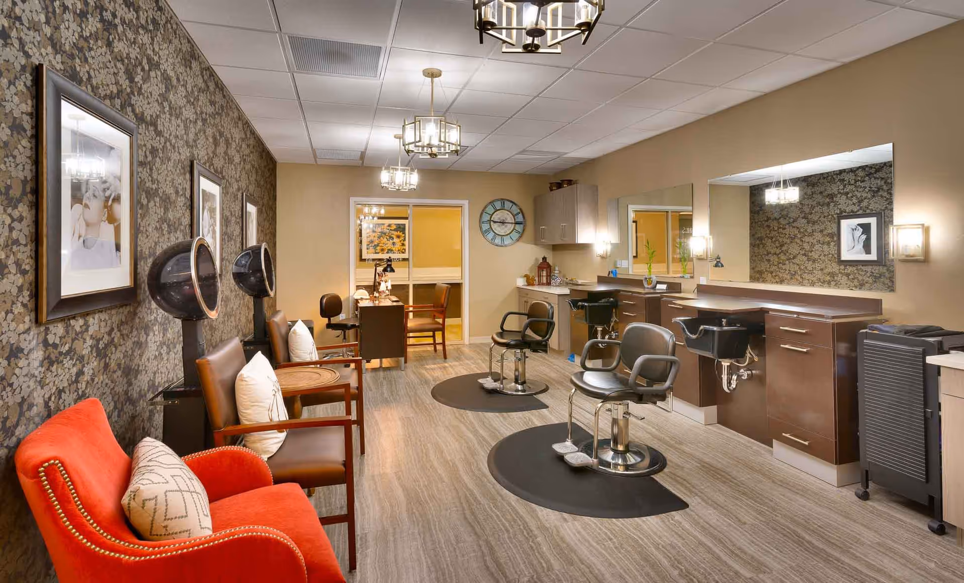 Interior view of a salon area in a senior living facility featuring two black salon chairs with footrests in front of a large mirror and sink stations. To the left, there are two brown chairs with white pillows under two black hair dryers, and a red armchair with a patterned pillow. The walls have floral wallpaper and beige paint, with framed black and white photos and a large clock. The floor has a light wood finish, and the room is lit by ceiling lights and wall sconces.