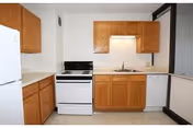 A small kitchen with wooden cabinets, a white refrigerator on the left, a white stove with an oven in the center, a sink with a faucet under a wooden cabinet, and a white dishwasher on the right.