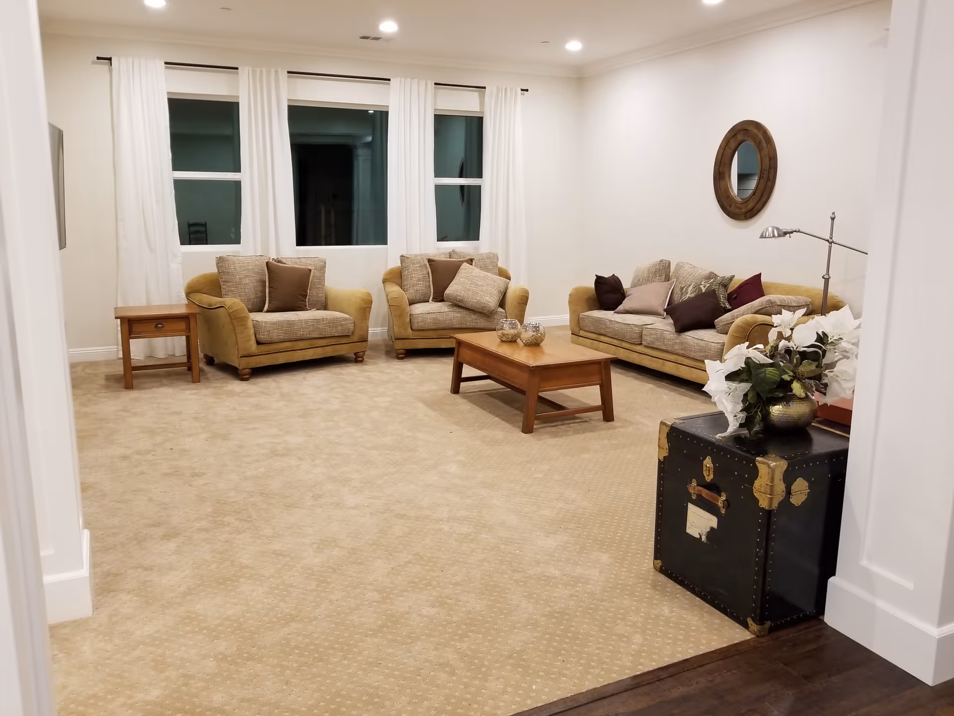 A cozy living room with beige carpet, two armchairs and a sofa with multiple cushions, a wooden coffee table, a side table, a round wall mirror, a floor lamp, and a decorative trunk with a vase of white flowers on top. The room has three windows with white curtains and recessed ceiling lights.