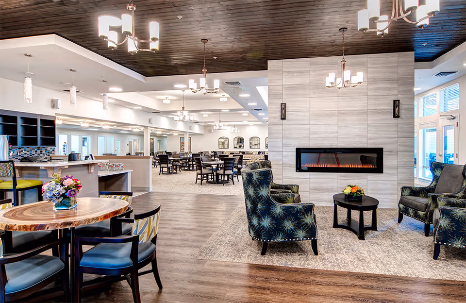 A spacious and well-lit senior living community common area featuring a modern electric fireplace set in a tiled wall. In front of the fireplace are three upholstered armchairs with a starburst pattern around a small round table with a floral arrangement. To the left, there is a wooden round table with chairs and a vase of flowers. The background shows a dining area with multiple tables and chairs, pendant lighting, and large windows allowing natural light to enter.