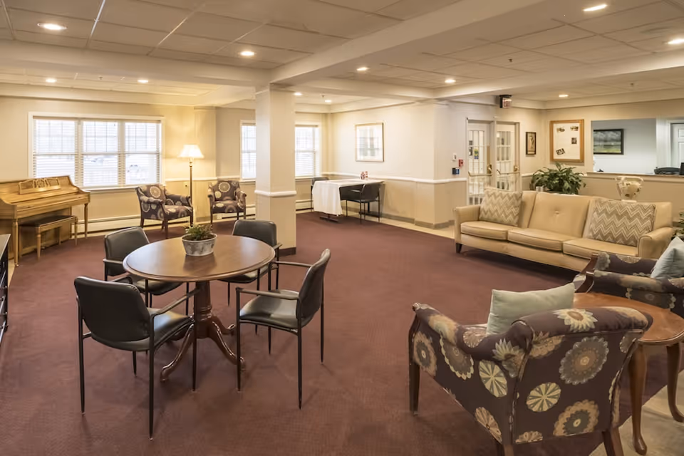 A bright senior living lounge with a round table and chairs, patterned armchairs, a beige sofa, and a piano by the windows.