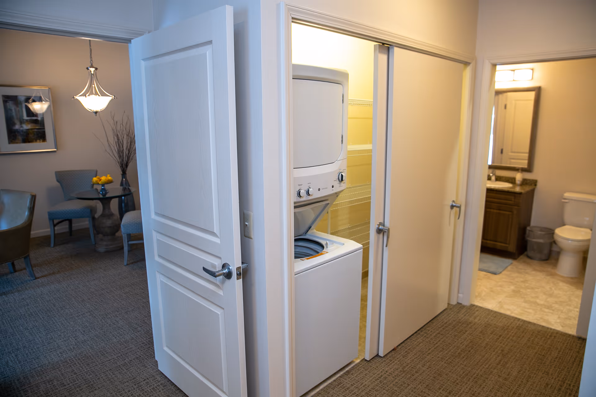 Interior view of a senior living facility showing a hallway with an open door revealing a stacked washer and dryer. To the right, there is a bathroom with a toilet, vanity with sink, mirror, and a trash bin. To the left, part of a dining or sitting area is visible with chairs, a table with a vase of yellow flowers, a framed picture on the wall, and a hanging light fixture.