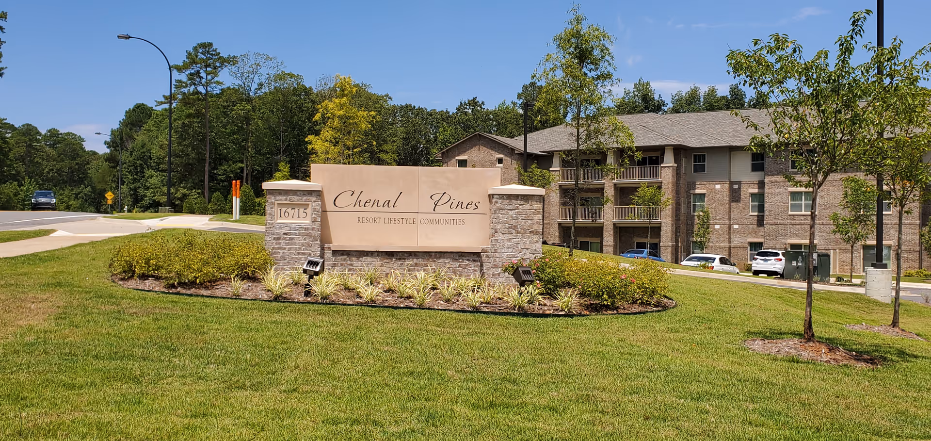 Outdoor view of the entrance sign for Chenal Pines Resort Lifestyle Communities with a landscaped area and a multi-story brick building in the background under a clear blue sky.