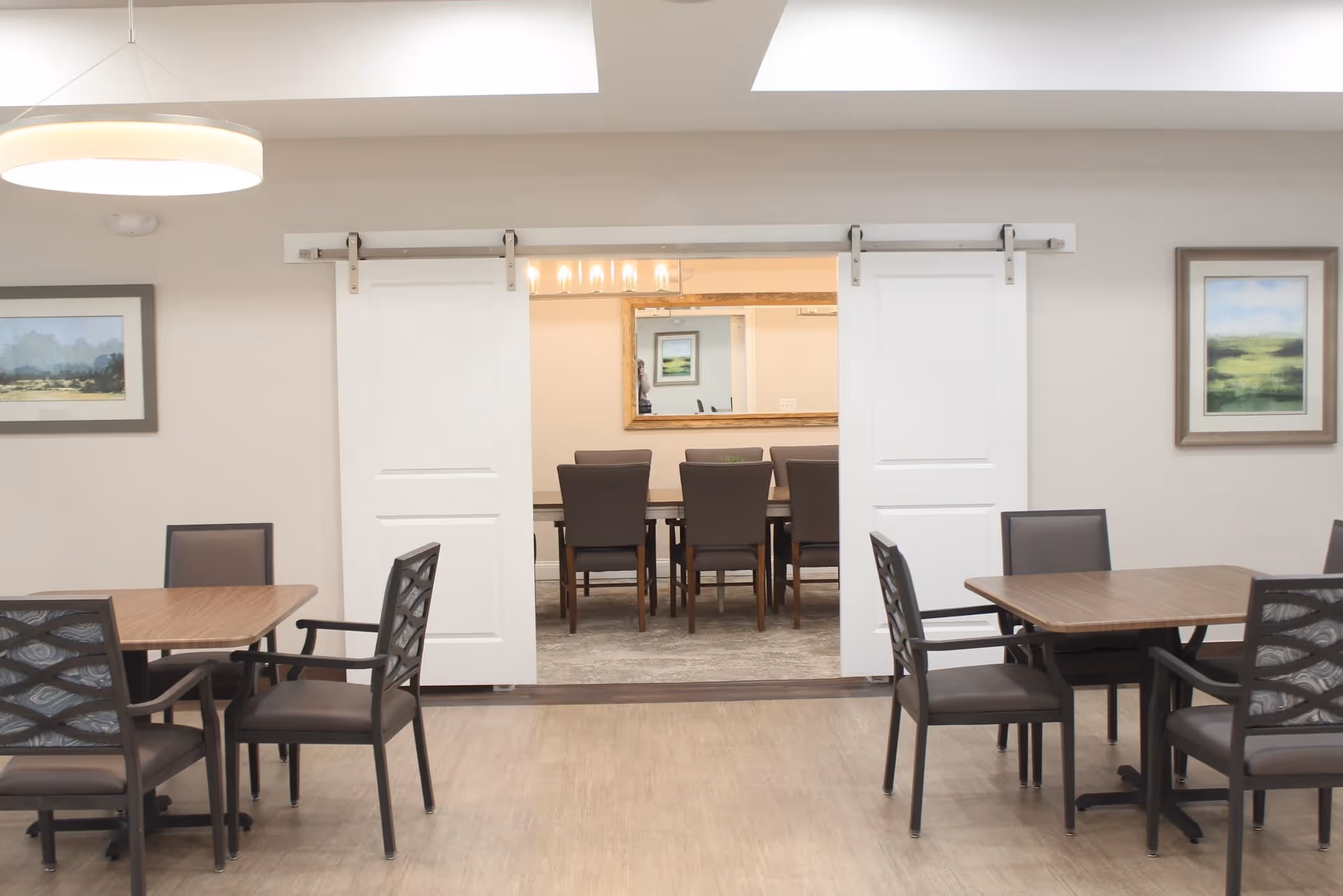 Interior view of a dining area with two square wooden tables and several chairs. In the background, there is a room with a long table and chairs visible through white sliding barn doors. The walls are decorated with framed landscape paintings, and a modern circular light fixture hangs from the ceiling.