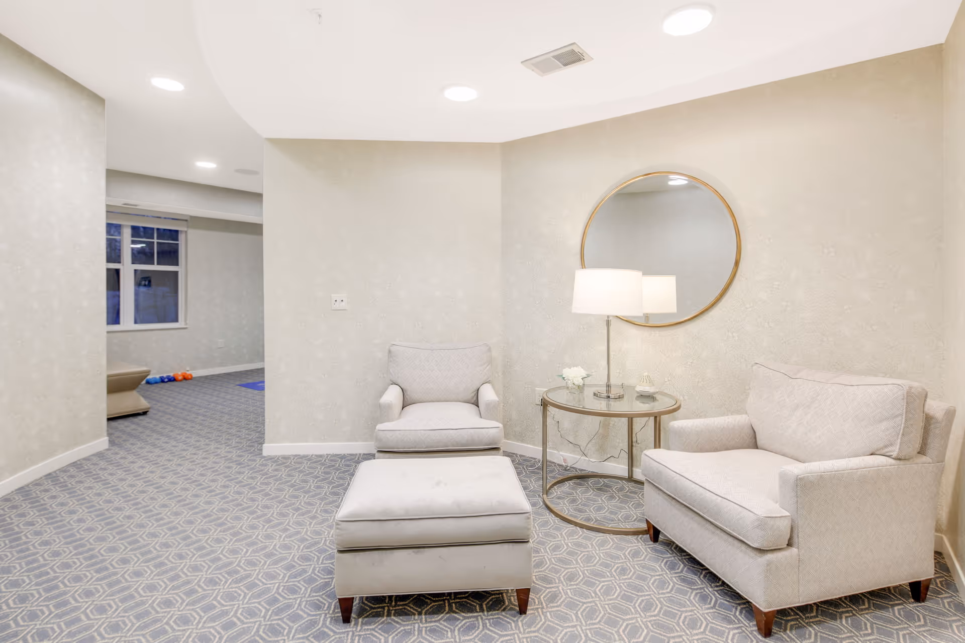 A cozy seating area in a senior living facility with a light gray armchair, a matching loveseat, and a cushioned ottoman. A round glass-top side table with a lamp and small decorative items sits between the chairs. A round mirror hangs on the wall above the table. The room has patterned carpet flooring and soft beige walls with recessed ceiling lights.