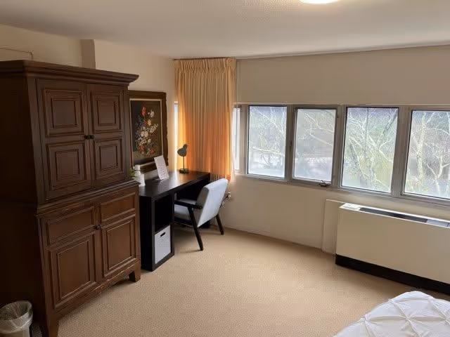 A bedroom with a large wooden armoire, a black desk with a white chair, a desk lamp, and a framed floral painting on the wall. There are four windows with beige curtains partially drawn, and a heating unit below the windows. The floor is carpeted and part of a bed with white bedding is visible in the foreground.
