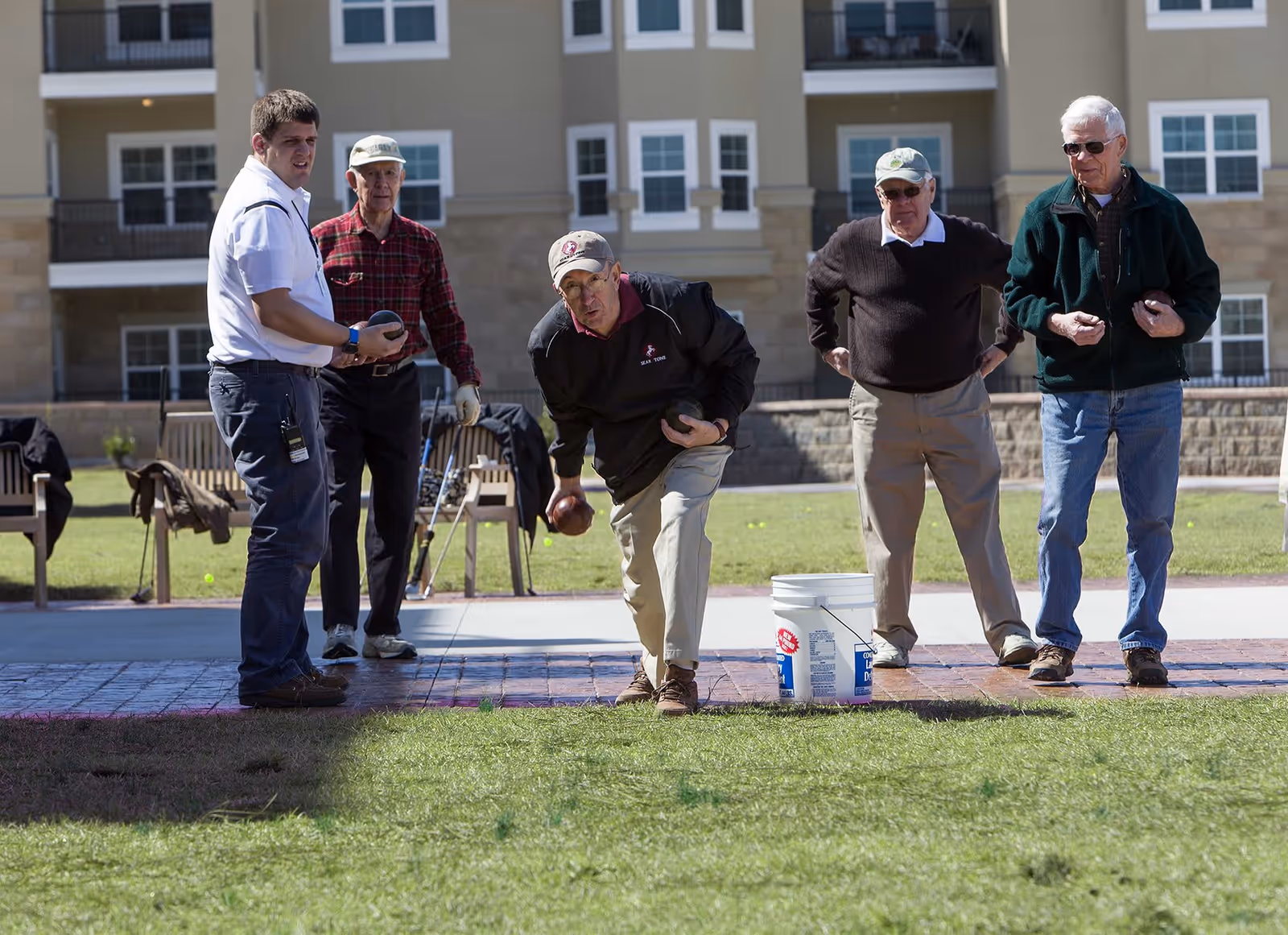 Five men outdoors playing bocce ball on a grassy area in front of a multi-story residential building. One man is in the process of throwing a bocce ball while the others watch. There are chairs and a white bucket nearby.