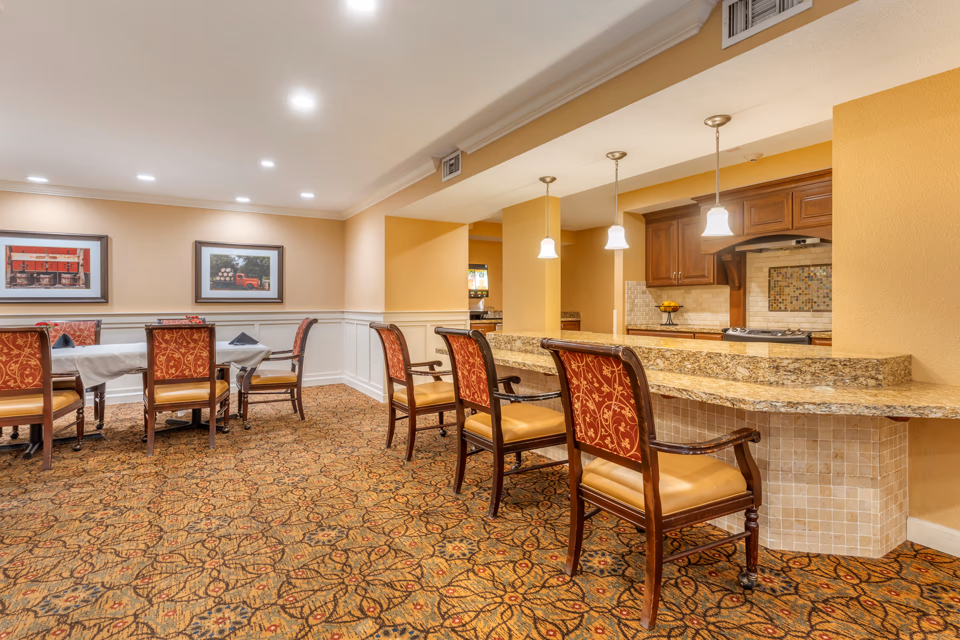 A dining area with a granite countertop bar and four ornate chairs with red and gold upholstery. Behind the bar is a kitchen area with wooden cabinets and a stove. To the left, there are two tables with chairs, each table covered with a white tablecloth and black napkins. The walls are painted beige with white wainscoting, and two framed pictures hang on the wall. The floor is covered with a patterned carpet.