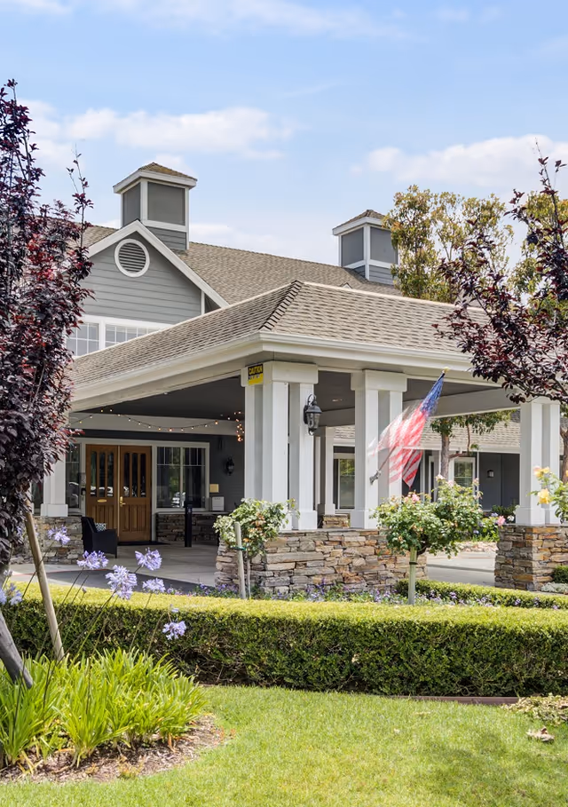 Exterior view of a senior living facility entrance with a covered driveway supported by white columns and stone bases. The building has gray siding, multiple windows, and two cupolas on the roof. There is an American flag hanging near the entrance, surrounded by well-maintained landscaping including green grass, bushes, and flowering plants.