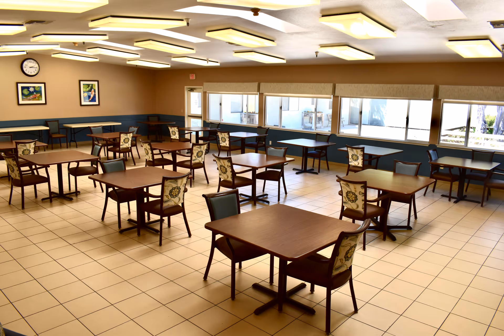 A spacious dining room with multiple square wooden tables and chairs arranged neatly. The chairs have floral patterned backs. The room has large windows along one wall letting in natural light, beige tiled floors, and ceiling lights. There are two framed pictures and a clock on one wall.