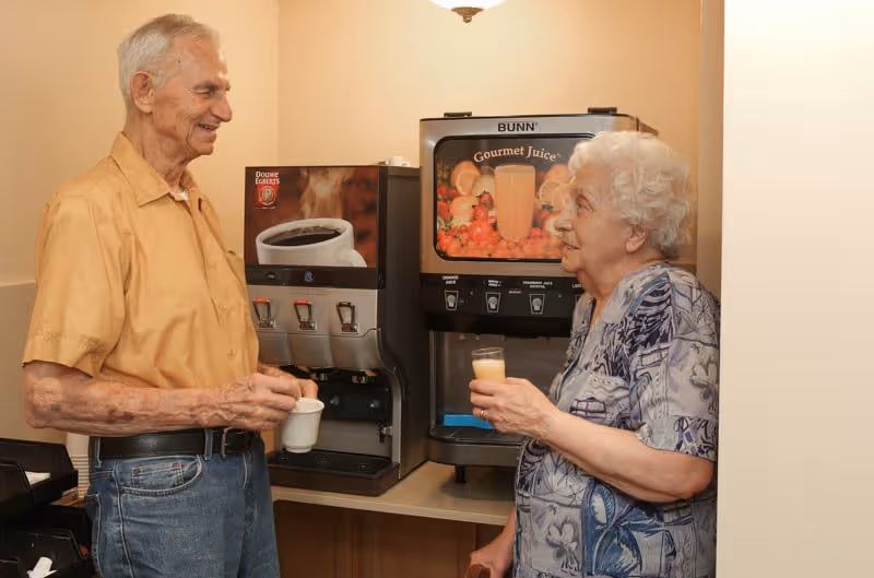 An elderly man and woman stand near beverage dispensers. The man is holding a cup of coffee, and the woman is holding a glass of juice. They are smiling and appear to be engaged in conversation in a well-lit indoor setting.
