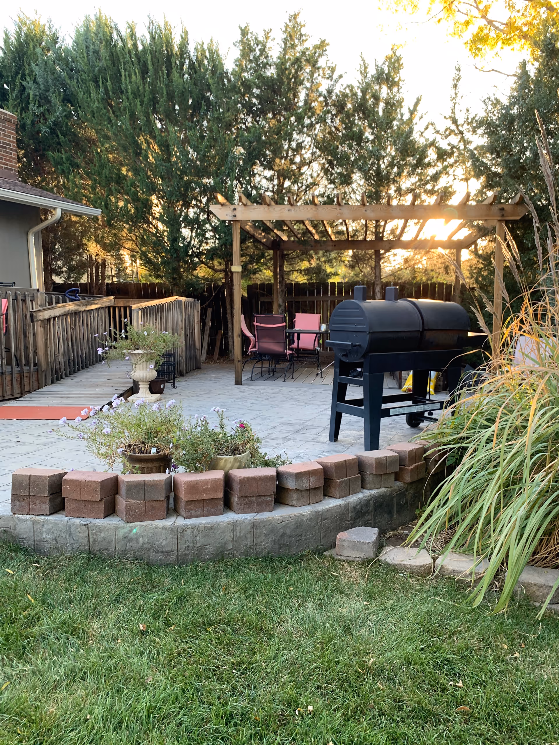Outdoor patio area with a wooden pergola, pink chairs around a table, a black barbecue grill, potted plants, and a grassy lawn in the foreground. Trees and a wooden fence are visible in the background with sunlight filtering through.