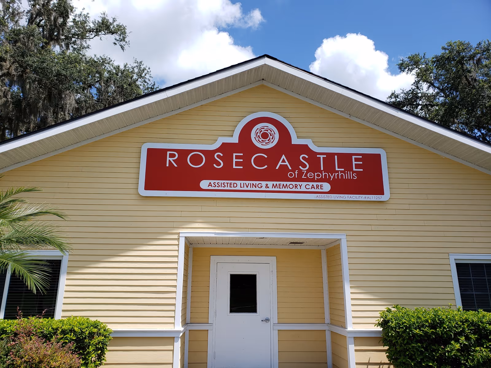 Front exterior view of a yellow building with a white door and two windows. Above the door is a large red and white sign that reads 'ROSECASTLE of Zephyrhills ASSISTED LIVING & MEMORY CARE'. There are green bushes and a palm tree in front of the building, with a partly cloudy blue sky in the background.