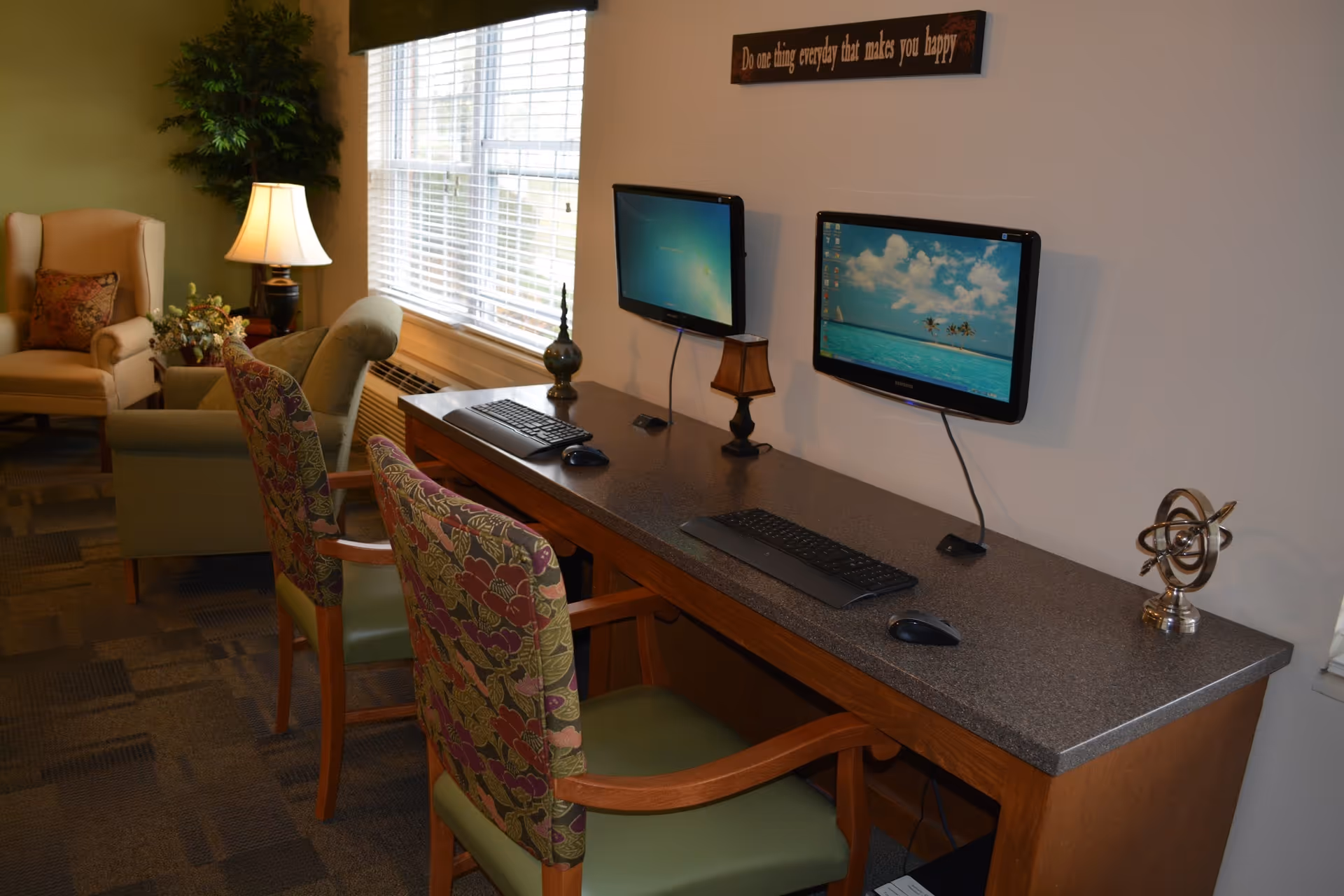 Communal computer workstation with two wall-mounted monitors, keyboards and floral-upholstered chairs in a cozy lounge area.