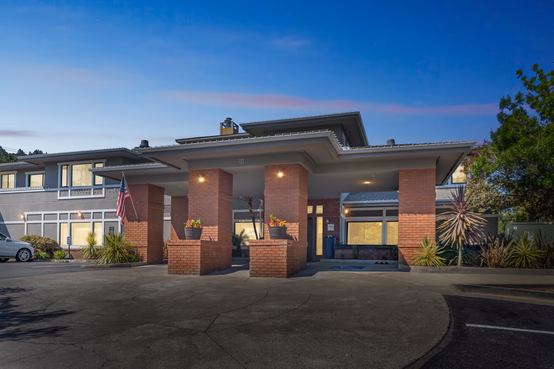 Front entrance of a two-story brick memory care building with a covered porte-cochere, planters, an American flag, and an evening sky.