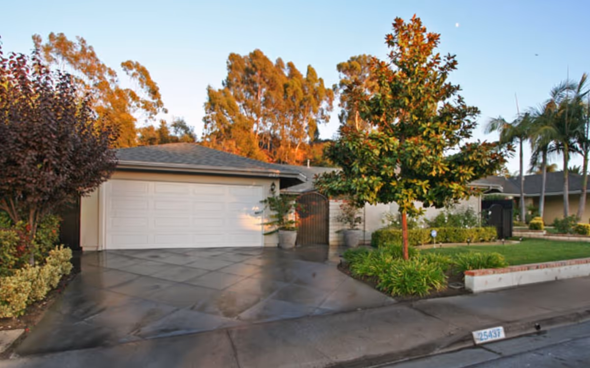 Exterior view of a single-story residential building with a white garage door, a driveway, and a well-maintained front yard featuring a tree, shrubs, and a lawn. The scene is set during daylight with clear skies and some tall trees in the background.