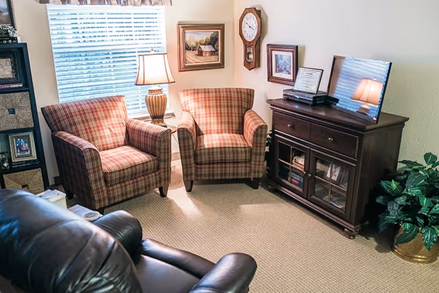 A cozy living room with two plaid armchairs and a dark leather recliner. A wooden cabinet with a flat-screen TV and framed pictures sits against the wall. A table lamp is placed between the armchairs near a window with blinds. The walls are decorated with framed artwork and a clock. A green potted plant is on the floor beside the cabinet.