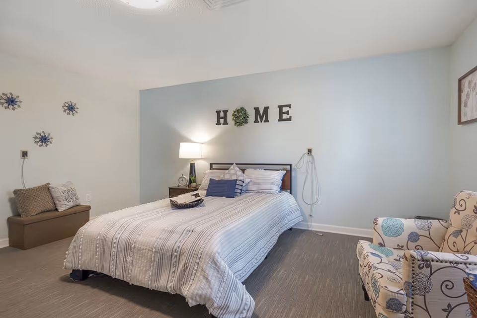 A light-blue bedroom with a double bed, 'HOME' letters on the wall, a bedside table with lamp, a patterned armchair and a bench.