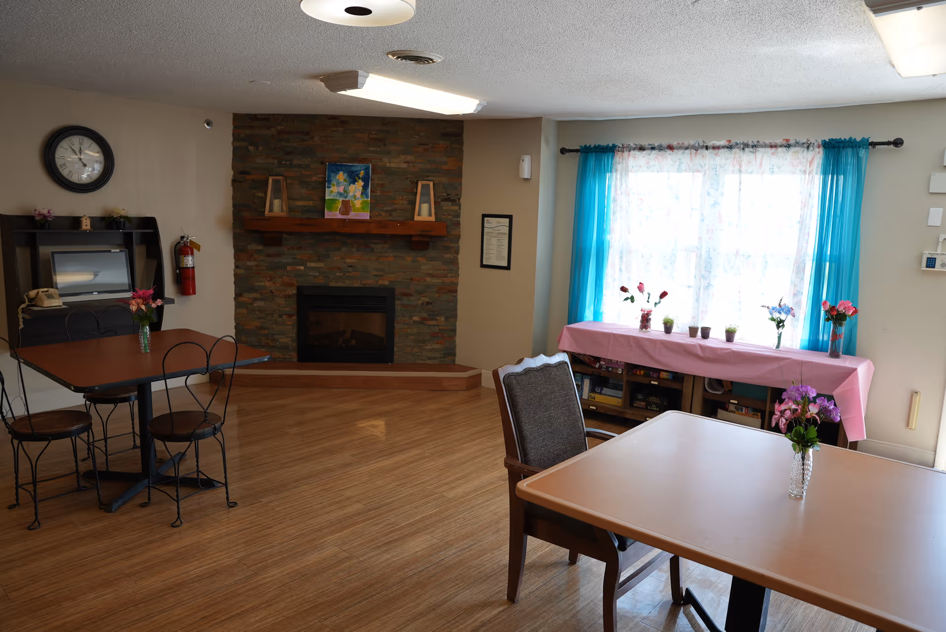 Bright common room with tables and chairs, a stone fireplace, and a window with turquoise curtains and floral decorations.