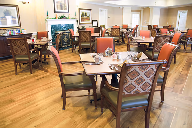 Spacious communal dining room with wooden floors, several set tables and upholstered upholstered chairs.