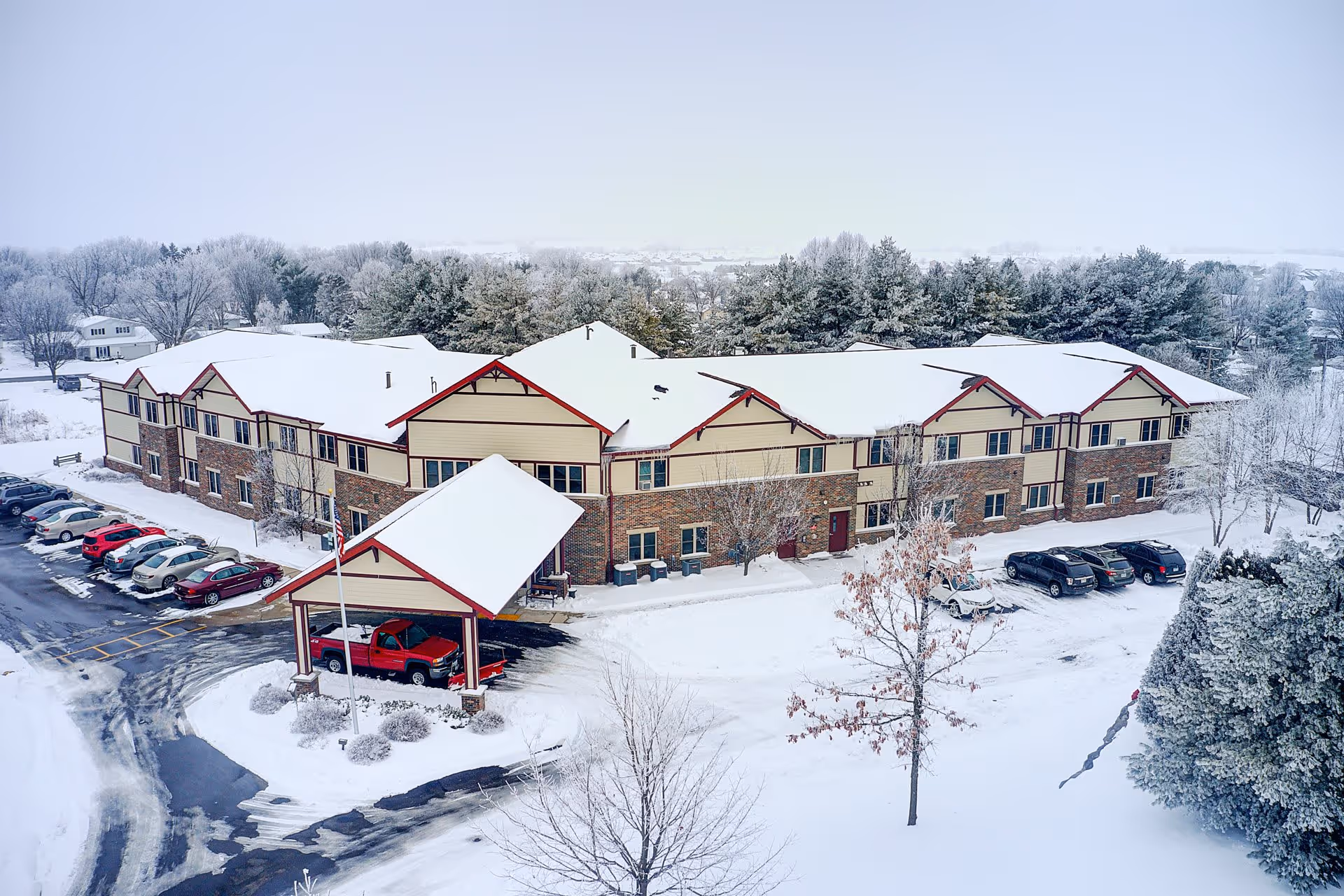 A large two-story senior living facility building covered with snow, surrounded by snow-covered trees and parked cars in a snowy parking lot. The building has a brick and beige exterior with red trim and a covered entrance with a red truck parked underneath. The landscape and roads are blanketed in snow, indicating a winter setting.
