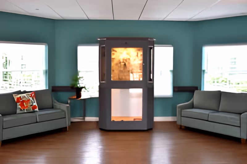 A living room with teal walls and wooden flooring featuring two gray sofas facing each other with a colorful pillow on the left sofa. Between the sofas is a small table with a potted plant. In the center against the wall is a modern fireplace with a glass front. Three large windows allow natural light into the room.