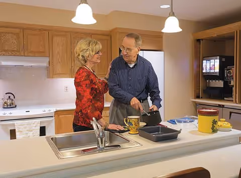 An elderly man and woman stand together in a kitchen. The man is holding a pot and appears to be cooking or preparing food, while the woman stands beside him smiling. The kitchen has wooden cabinets, a white stove, a sink in the foreground, and a beverage dispenser on the right.