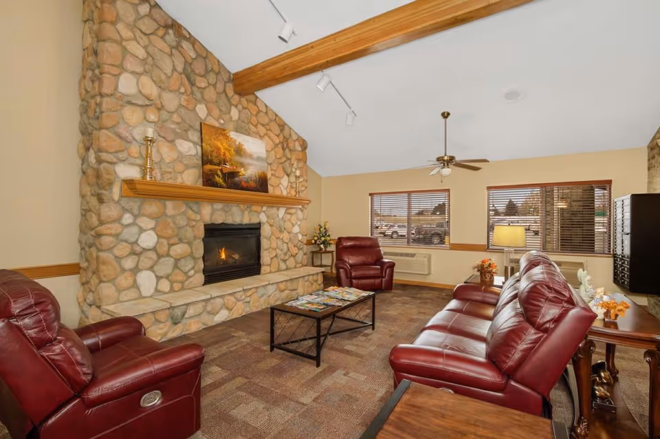 A cozy living room with a stone fireplace, a wooden mantel holding decorative items and a painting above it. The room features red leather recliners and a matching sofa arranged around a coffee table with magazines. There are two windows with wooden blinds, a ceiling fan, and a table lamp providing warm lighting.