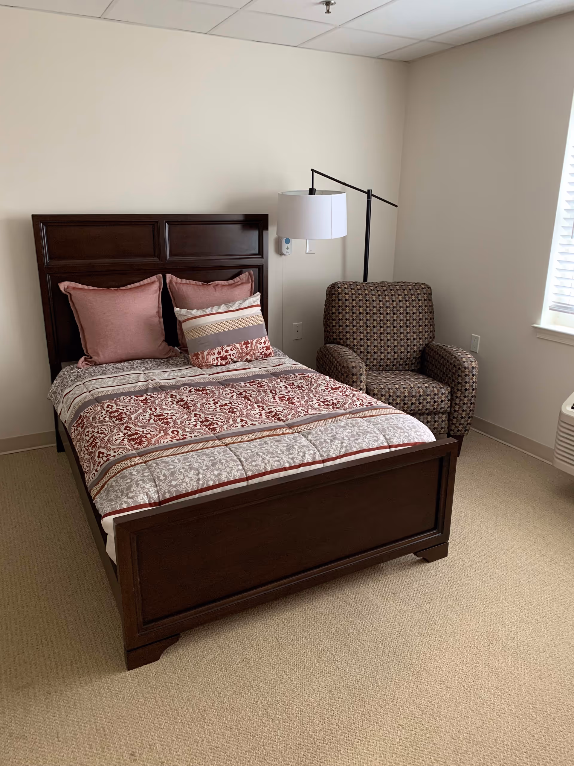 A bedroom with a dark wooden bed frame, a bed made with patterned bedding in shades of red, gray, and white, and three pillows. Next to the bed is a patterned armchair and a floor lamp with a white lampshade. The room has beige carpet and a window with white blinds.